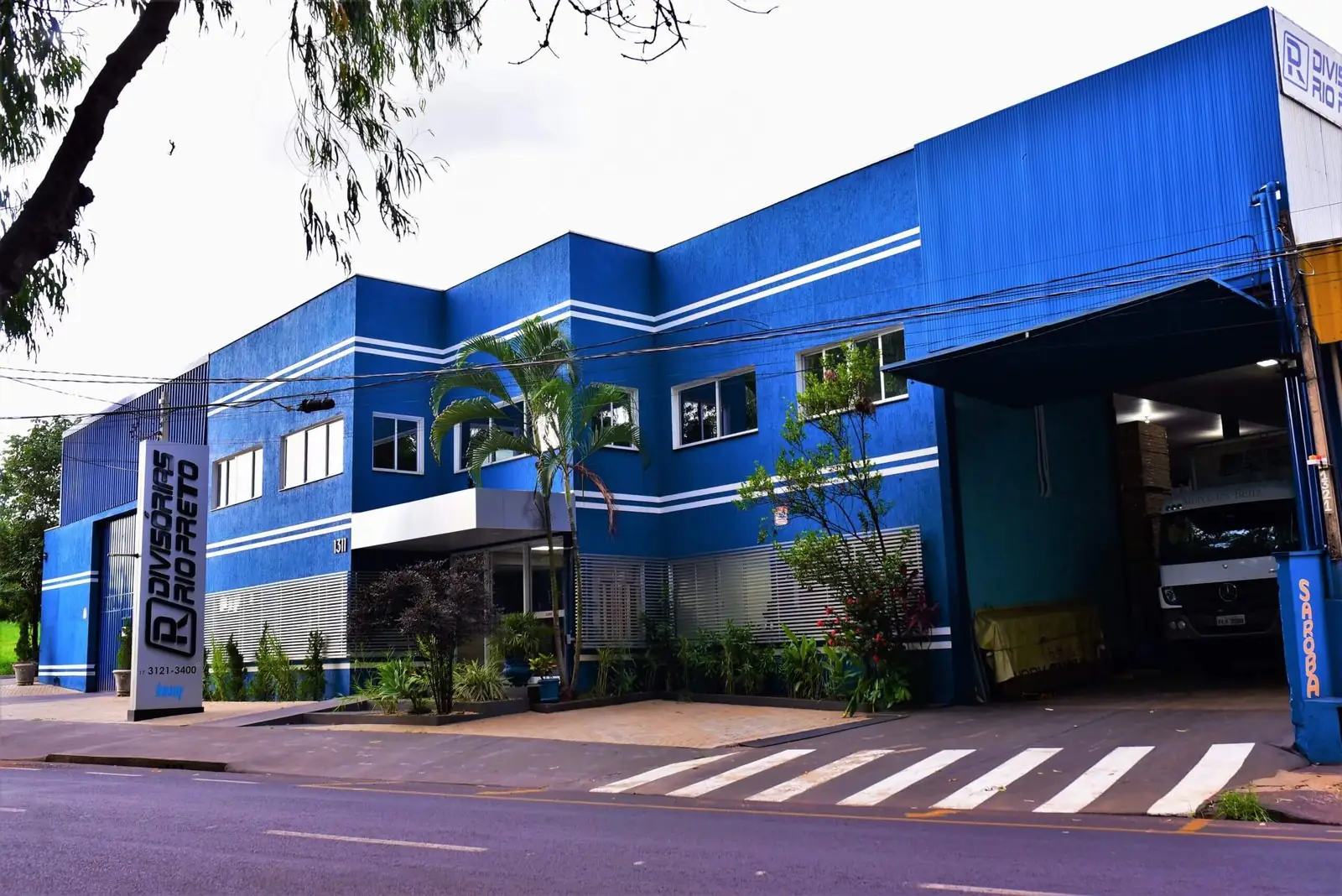 Fachada azul de dois andares da Divisórias Rio Preto na Av. Paulo de Oliveira e Silva, com o totem da marca na calçada e o galpão de carga à direita