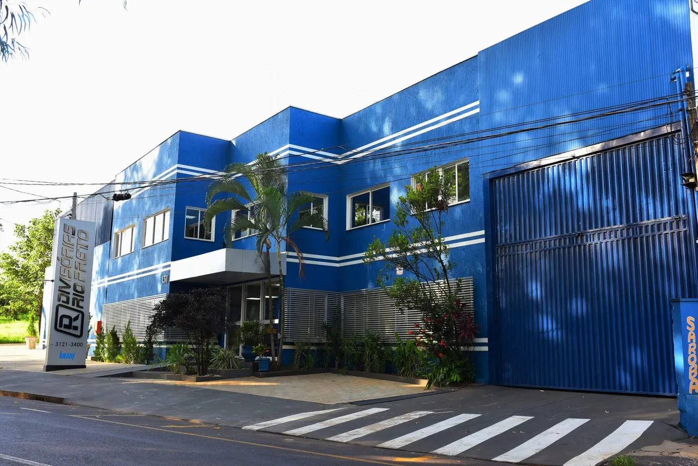 Fachada azul de dois andares da Divisórias Rio Preto na Av. Paulo de Oliveira e Silva, com o totem da marca na calçada e o galpão de carga à direita